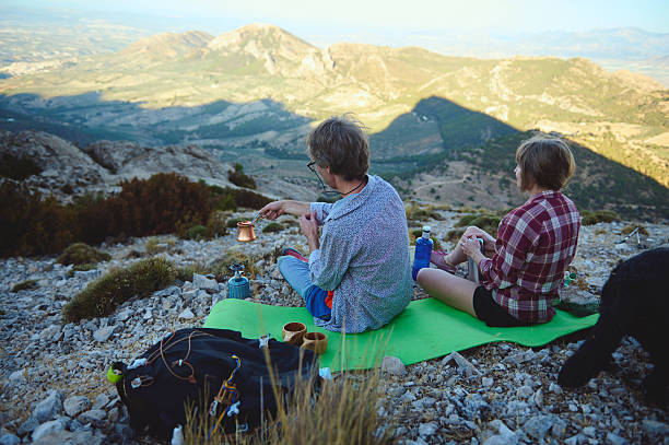 A pair relaxes on a mountain, enjoying drinks from a camping setup surrounded by nature. The clear blue sky and vast rolling plains create a stunning backdrop, perfect for outdoor adventures.
