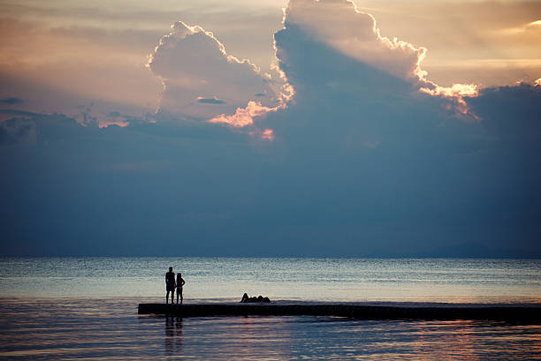 Koh Phangan, Thailand - February 4, 2017: Breathtaking sea view at sunrise. Silhouettes of unrecognizable tourists standing on pier. Gray water surface with small ripples, cloudy sky with sunbeams