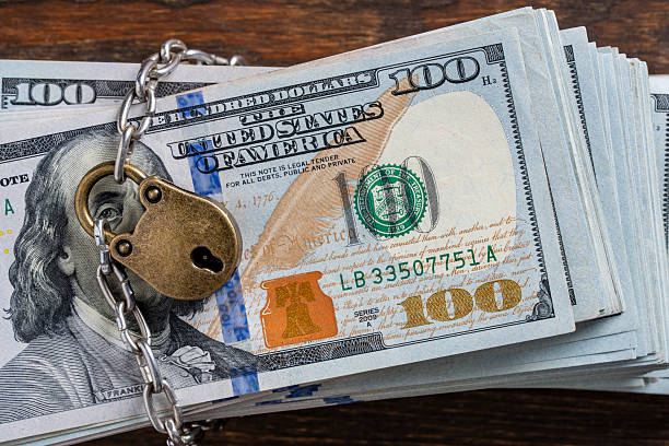 istock Metallic chain and padlock around american one hundred dollar banknotes, closeup, top view. Business safety or financial protection or restriction access. Heap of money in chain with padlock 2236139737