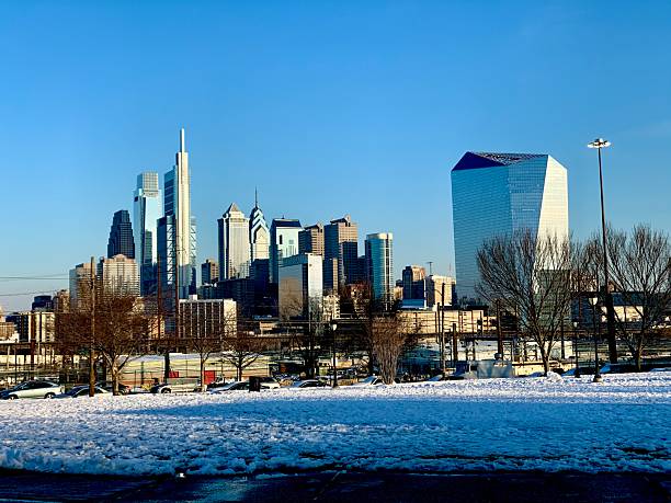 Sleek modern skyscrapers tower above the snow-covered ground in downtown Philadelphia, set against a crisp, clear blue winter sky. The scene highlights the city's iconic skyline in a serene seasonal setting
