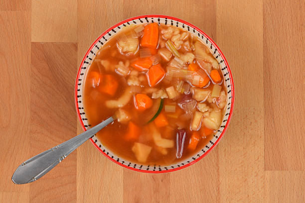 A bowl of hot vegetable and pasta soup with a spoon, placed on a wooden surface