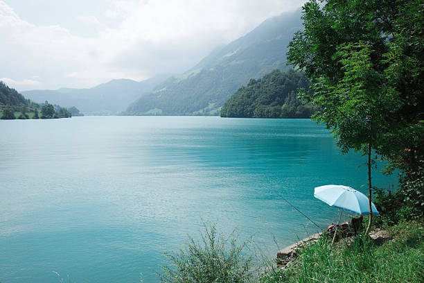 The turquoise water is surrounded by lush greenery and forested hills. A blue umbrella provides shade over a small seating area near the shore.