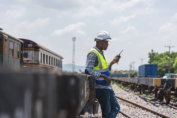 istock Railway Worker Using a Walkie-Talkie at a Train Station, African American Engineer Communicating on a Two-Way Radio at a Rail Yard, Foreman in a Hard Hat and Vest Standing by Train Cars 2236185639