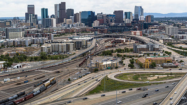 Elevated view of transportation infrastructure in Denver, Colorado showing multiple highways, railway tracks, and freight trains alongside urban development. The foreground displays active rail yards with cargo trains while interstate highways with flowing traffic curve through the area. Mid-ground features residential and commercial buildings of Five Points neighborhood. Background showcases Denver's downtown skyline with high-rise office buildings against distant mountain backdrop. Bird's eye view captures the interconnection between transportation systems and urban development.