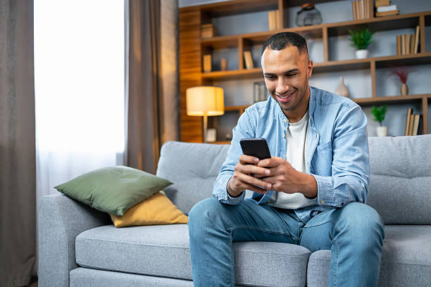 istock Handsome african american man using smartphone while relaxing sitting  on couch at home in living room, browsing internet or messaging with friends, Free Copy Space 2236200651