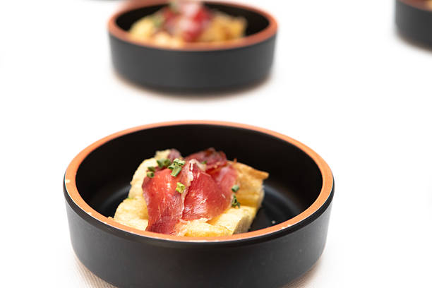 Small elegant black bowls containing delicious appetizers with cured ham and bread are arranged on a white surface, ready for a catering event