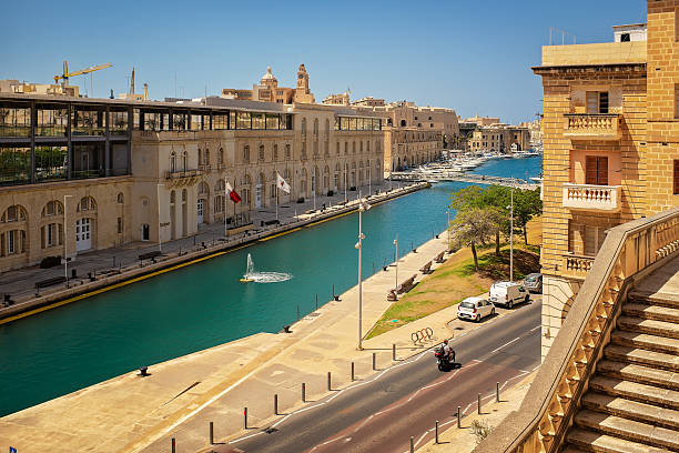 Bormla, Malta - 07/13/2025: Scenic view of a canal with a park, city road, stairs, and a bridge framed by historic Maltese architecture under the summer sun.