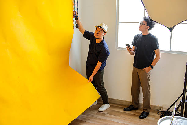 Photographer Adjusts Vibrant Yellow Backdrop As Model Observes With Phone In Hand
