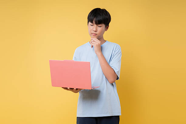 istock Teenage boy thoughtfully uses a laptop against a bright yellow backdrop; concentration and tech 2236218308