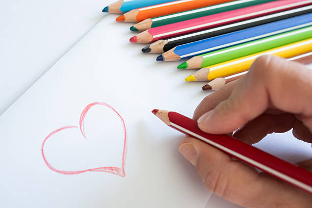 A hand holds a vibrant red pencil above an envelope adorned with a heart, with softly blurred colorful pencils in the background. Process of crafting personal Valentine's messages and handmade cards.