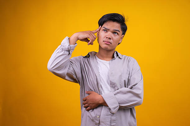Young Asian Man in Casual Tshirt Showing Thinking Gesture Aginst Yellow Studio Background