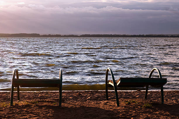 Two empty benches sit on the sandy shore, facing a tranquil lake. Soft waves ripple under warm golden sunlight as clouds drift lazily above.