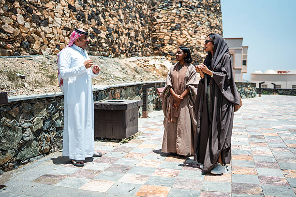 Mid adult Arabic females engaging with an Arabic tour guide at Shamsan Ottoman Castle featuring historical stone architecture and traditional attire under clear blue skies. The group converses and enjoys the cultural experience.