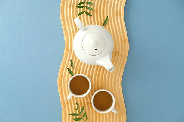 istock Minimalist tea composition with white teapot and cups on wavy wooden surface over blue background, accented with green leaves for a fresh, modern look 2236227402