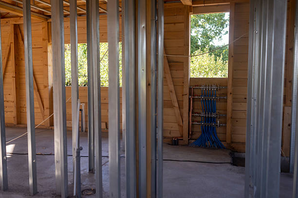 A partially constructed wooden house interior featuring exposed metal frames, wooden walls, and a visible plumbing system. Bright daylight illuminates the space through open window frames.