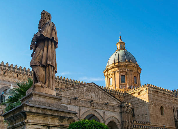 Saint Jerome statue outside Palermo Cathedral in Sicily, Italy. Background: cathedral's 18th century Baroque cupola of oxidized lead sheeting atop a drum with rectangular windows framed by pilasters & triangular pediments. 12th century Norman walls feature blind arches, interlaced arcading & crenellations at the roof. 15th century South Portico with pointed arches and a sculpted tympanum above with a relief of the Madonna & Child. Unesco listed