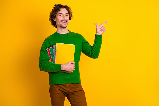 istock Smiling man holding colorful notebooks on a vibrant yellow background while gesturing with his finger and wearing green 2236250674