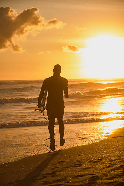 The picture shows a foggy early morning scene at Huntington Beach. A person is walking along the sandy beach, carrying a blue surfboard. The fog obscures the background, creating a hazy, dreamlike atmosphere.