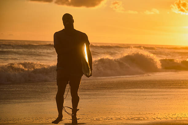 Silhouette of unrecognizable surfer sit on the sand along the shoreline, with waves crashing in the background, during a breathtaking sunset