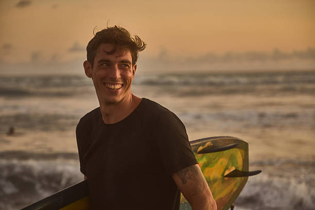 istock Smiling surfer holding board while standing on beach at sunset 2236251804