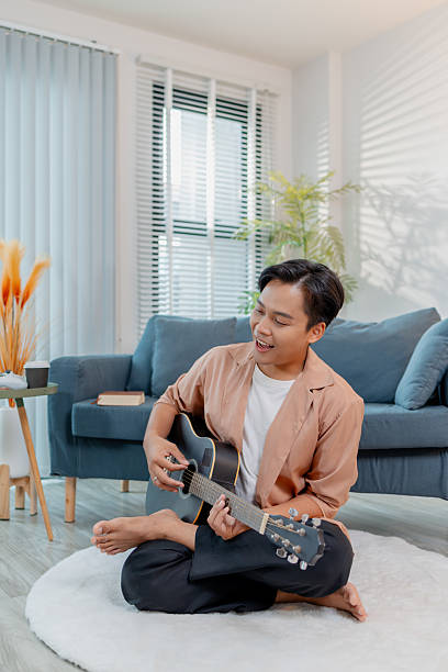 istock A young man sits cross-legged on the floor at home, joyfully playing an acoustic guitar while singing. His relaxed posture and happy expression reflect creativity and passion. 2236254570