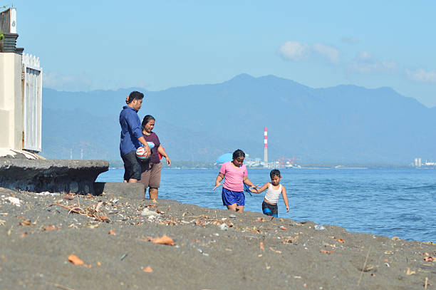 A family walks together, parents carrying supplies along the villa wall while the children stroll nearby, looking down toward the sandy surf waters.