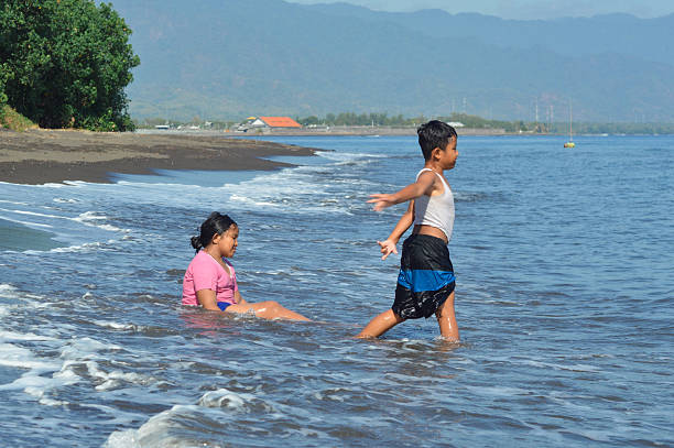 A girl sits wet on the shore while a boy steps toward the surf, both enjoying the lively splash of seawater along the sandy beach.