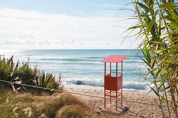 Red lifeguard hut standing on a quiet sandy beach, framed by green plants and a rope barrier, overlooking blue ocean water with gentle waves under a bright sky