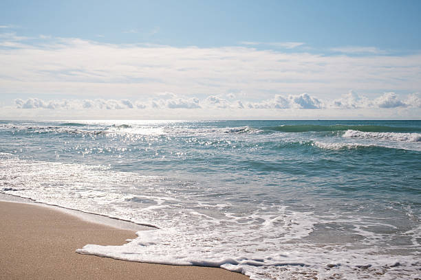 Ocean waves are breaking on a sandy beach under a bright blue sky with scattered clouds, creating a peaceful and serene natural landscape with sun glistening on the water surface