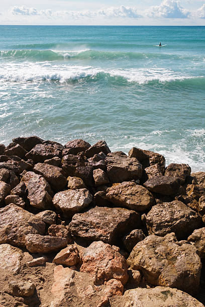 Surfer sitting on a surfboard in the vast blue ocean, waiting for the perfect wave, with rugged rocks forming a natural barrier on the foreground under a bright sky