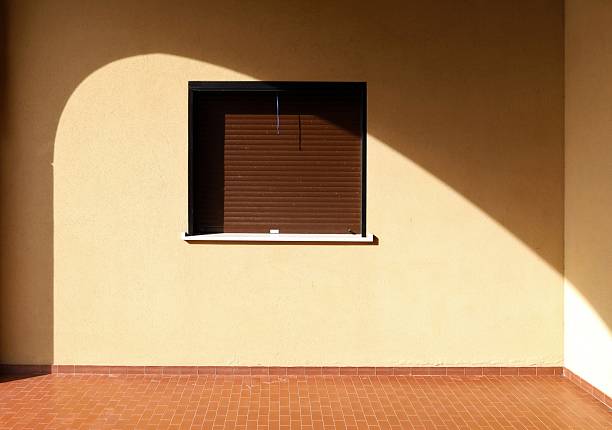 Closed brown shutter on yellow plaster wall with a shadow of an arch around it. Tiles eartenware floor. Background for copy space.