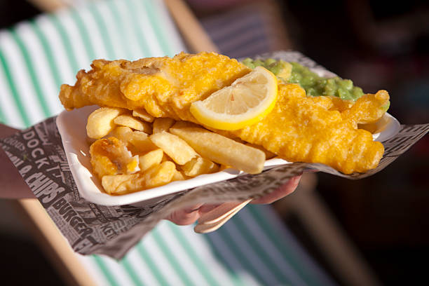Hand holding fish and chips with mushy peas and a slice of lemon on a card tray