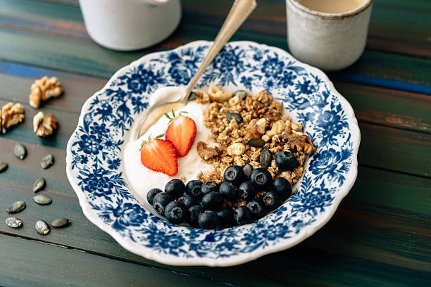 Ein blauer Teller, gefüllt mit Granola, Nüssen, Joghurt, Erdbeeren und Heidelbeeren. Daneben steht ein Kännchen mit Milch und eine Tasse mit Kaffee.