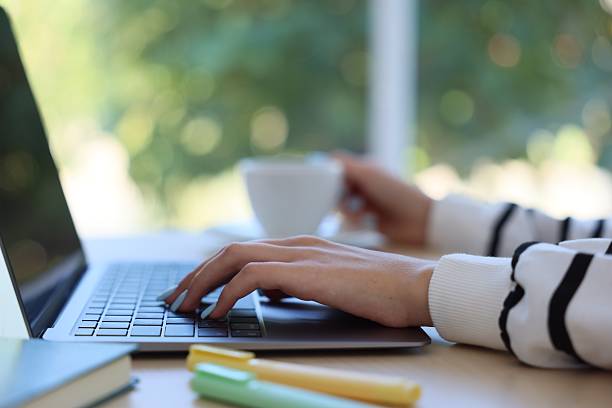 istock Woman with coffee working on laptop at table against blurred background, closeup 2236278878