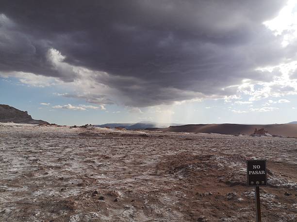 A dramatic view of Valle de la Luna in the Atacama Desert, Chile, featuring red rock, salt flats, and a ‘No Passing’ sign under stormy, menacing clouds framed by blue sky. This scenic, arid landscape showcases the natural beauty and moody atmosphere of the high-altitude Altiplano region.