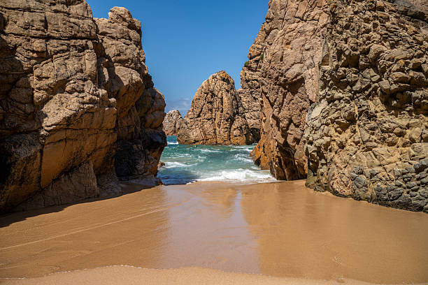 Turquoise waves crash against dramatic rock formations at a secluded sandy cove in Praia da Ursa, near Cabo da Roca, Portugal’s rugged Atlantic coastline.