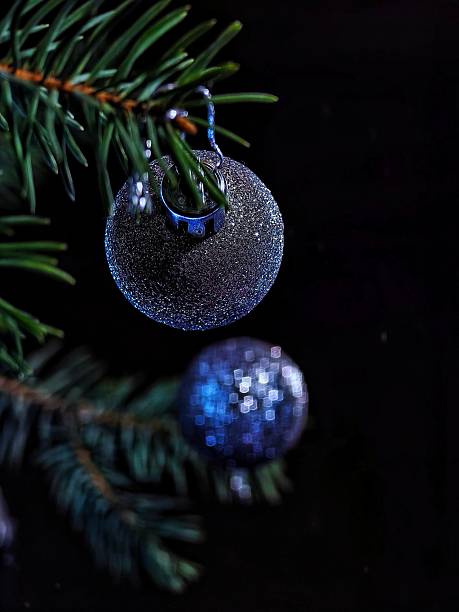 Sparkling silver-blue Christmas bauble hanging on a green fir branch against a dark background. Festive atmosphere and winter coziness.
