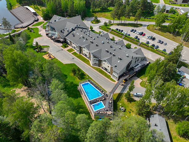 Suzdal, Russia - May 12, 2024: An aerial view of a modern hotel building with a long, grey roof, a swimming pool with two people swimming, and a lake in the background.