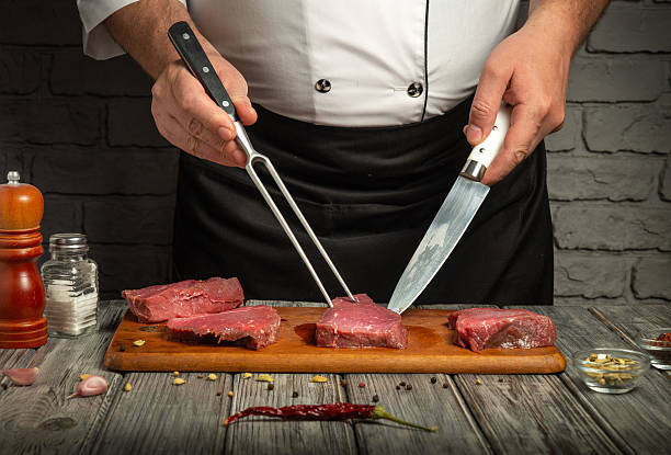 istock A chef stands in a cozy kitchen, using a fork and knife to precisely cut juicy beef steaks on a wooden board. Aromatic spices and herbs are scattered nearby, enhancing the culinary atmosphere 2236292531