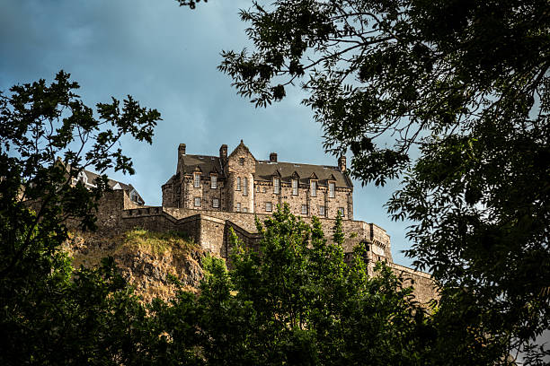 Edinburgh, Scotland - June 29, 2022: View of Edinburgh Castle in Scotland, perched on Castle Rock and framed by green trees in the foreground. The historic fortress stands prominently against a cloudy blue sky, showcasing its medieval stone architecture.