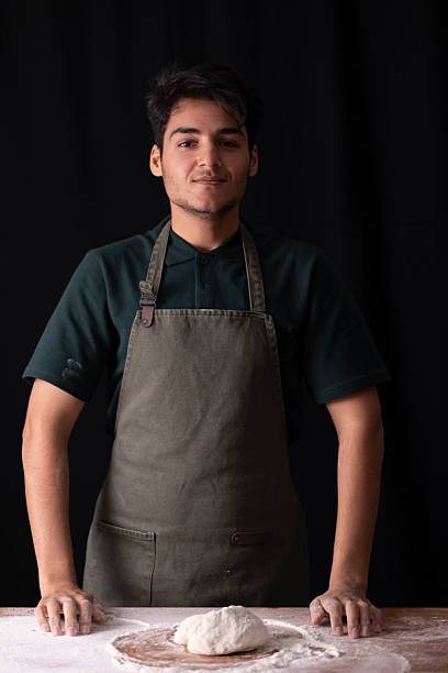 Young man in an apron standing with dough and flour, preparing food on a wooden table