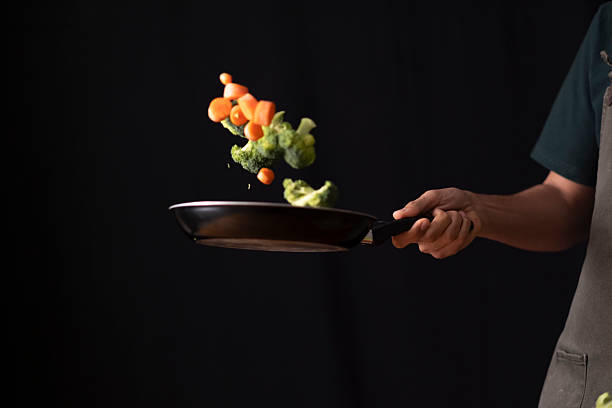Cook's hand holding a frying pan, tossing vegetables in motion against a black background