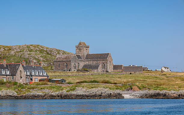 A scenic coastal view of Iona Abbey and the adjacent Nunnery on the island of Iona, Scotland. Captured from the sea in August, the image features historic stone architecture surrounded by grassy terrain, rocky shoreline, and calm blue waters. The background includes traditional houses with chimneys and rolling hills under a clear sky. This peaceful setting evokes spirituality, history, and timeless beauty. Ideal for editorial and commercial use in themes such as religious heritage, cultural tourism, and historical architecture. The image highlights the serene character of the Scottish islands and their rich cultural legacy.