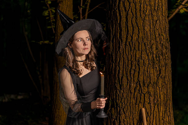 Woman in witch costume holding candle by tree in dark forest with spooky halloween night mood