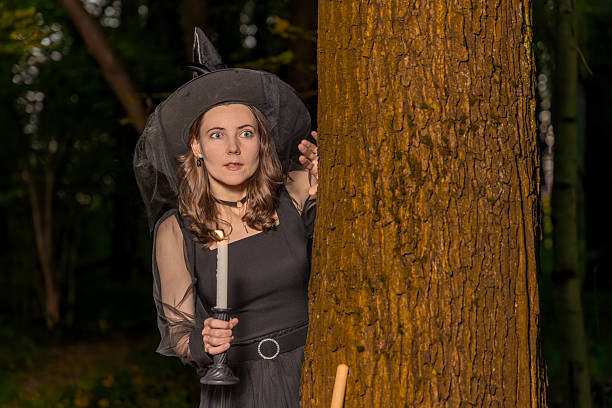 Woman in witch costume holding candle peeks from behind tree in dark forest at night with spooky halloween mood