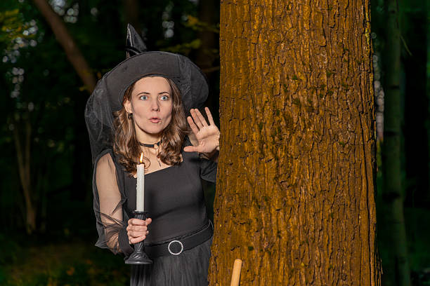 Woman in witch costume holding candle makes shh gesture behind tree in dark forest at night with spooky halloween mood