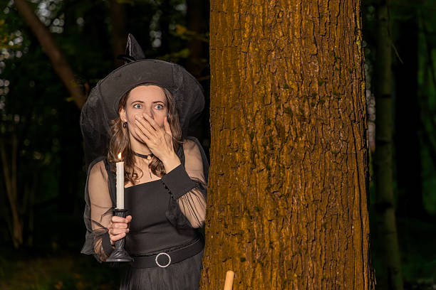 Woman in witch costume holds candle and covers mouth behind tree in a dark forest at night with spooky halloween mood