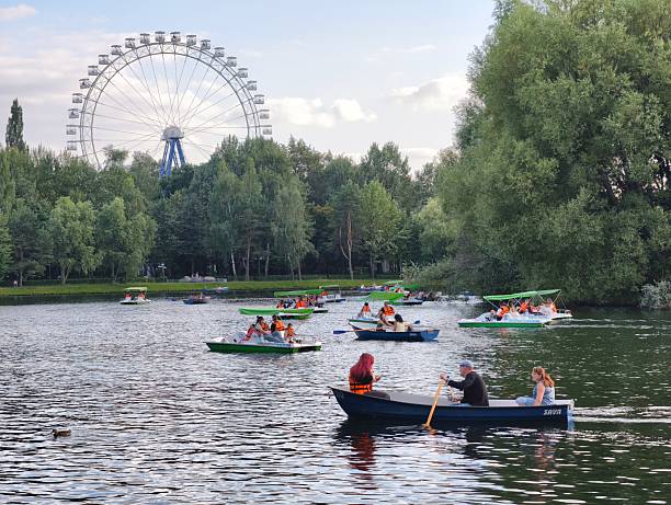 Moscow, Russia - September 07, 2025: This image captures a lively summer day at the pond in Izmaylovsky Park. Visitors enjoying paddle boats and rowboats with a large Ferris wheel visible in the background