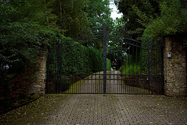Elegant wrought iron gate with ornate details between stone pillars, leading to a private driveway surrounded by lush greenery.