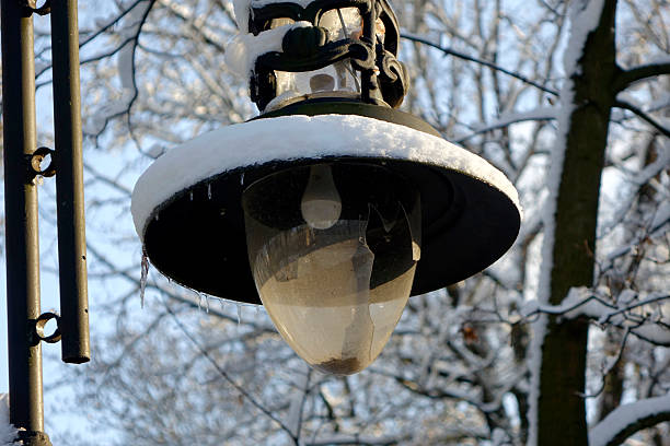An old lantern with shattered glass hangs in a snowy park, surrounded by bare trees glistening under the winter sun.